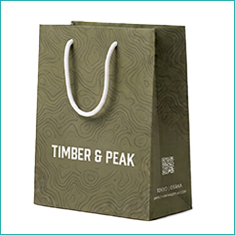 男性向けブランドのショッパー。等高線をモチーフにしたカーキ色の背景に、白字で「TIMBER＆PEAK」のロゴ。サイドにはQRコードがプリントされ、紐は白のアクリル丸紐