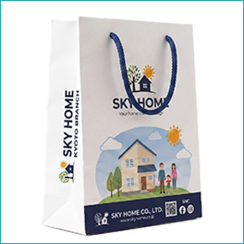 住宅展示場の資料配布用紙袋。一軒家と家族のイラストに「SKY HOME」のロゴ、QRコードやSNSアイコンを配置。サイドにもロゴのプリントあり