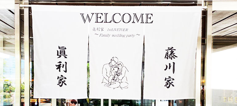 白色生地の3巾のれんの、左右両端の巾に黒い文字で、新郎新婦両家の苗字が名入れされていて、真ん中の巾には新郎新婦とお子さんのイメージイラストと「WELCOME」の文字が名入れプリントされた、結婚式用の3巾のオリジナルウェルカムのれんの実例画像